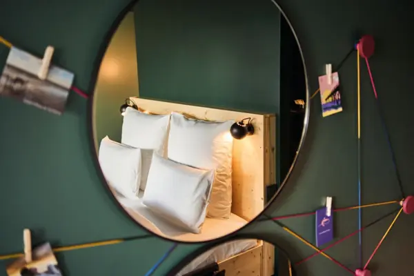 Zimmer im Urban Nature Hotel St. Peter-Ording In einem Spiegel an einer grünen Wand ist ein gemütliches Bett zu sehen mit weißer Bettwäsche und vier Kissen.