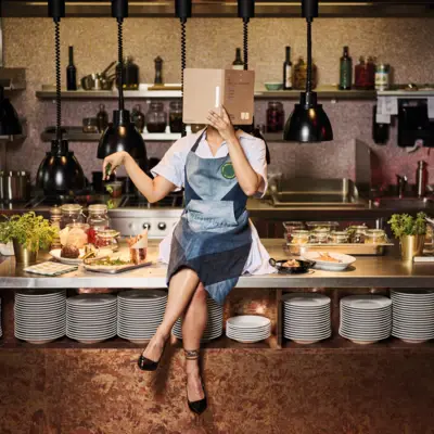 Speisekarte Auntie Clara Eine Frau in Schürze sitzt auf einer Küchenarbeitsplatte und hält eine Speisekarte vor dem Gesicht.