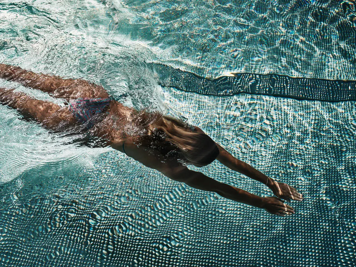 Alpine Swim Club Badeschloss Eine Frau taucht durch einen hellblauen Pool.