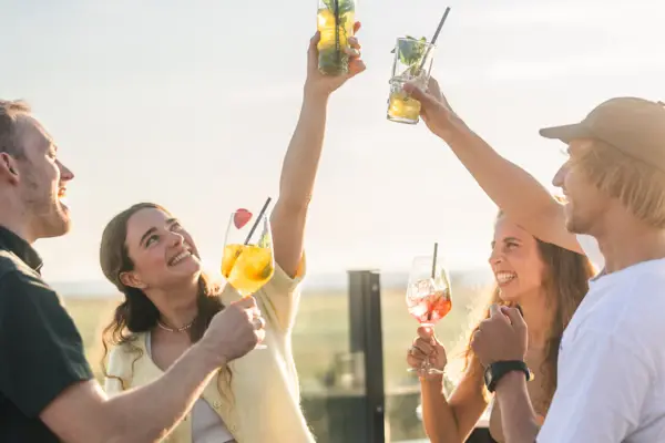 Scraper’s Club People Die Rooftopbar Scraper's Club im Urban Nature Hotel St. Peter Ording mit 4 Personen, die bei Sonnenschein mit Drinks anstoßen und die Drinks hochhalten..