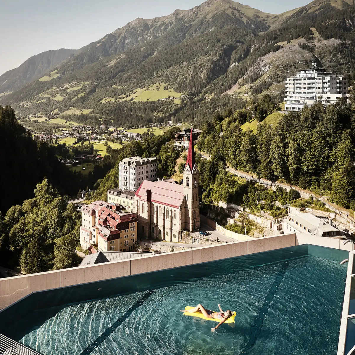 Schwimmen mit Aussicht – Sommergenuss über den Dächern von Bad Gastein Infinity Pool mit Ausblick auf Bad Gastein und die Alpenlandschaft – Frau entspannt auf gelber Luftmatratze mit Blick auf Kirche und historische Gebäude