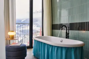 Badewanne Badewanne mit blauem Rock in einem Badezimmer mit Fenster, das auf eine verschneite Bergkette blickt.