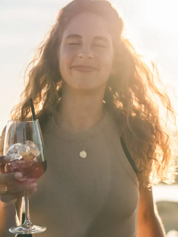 Drinks Scraper’s Club Eine Frau lächelt und hält einen Drink in einem Weinglas in ihrer Hand, hinter ihr scheint die Sonne.