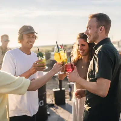 Scrapers Club St. Peter-Ording Eine Gruppe von Menschen steht auf der Rooftop Bar Scraper's Club St. Peter-Ording mit Getränken in der Hand und mit Blick auf die Salzwiesen und das Meer.