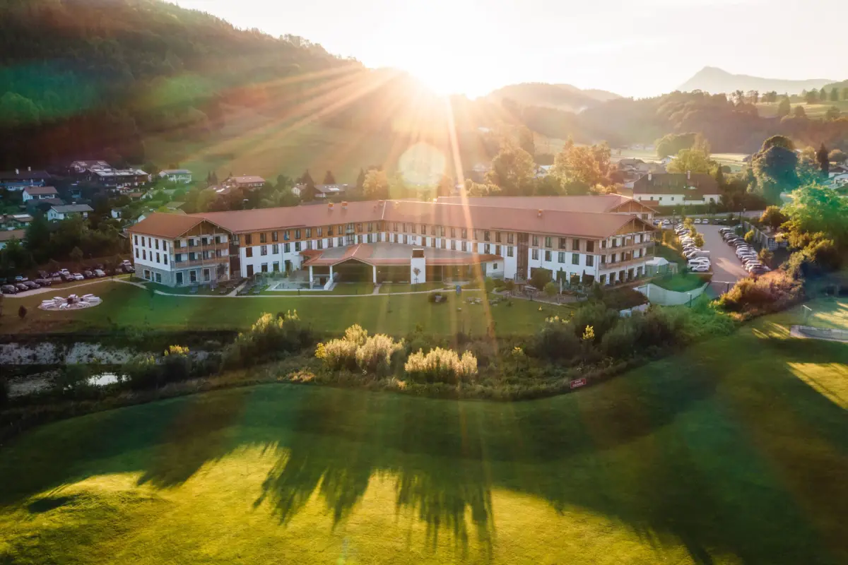 aja Hotel in Ruhpolding Luftaufnahme eines großen Hotels mit weitläufigem Garten in der Alpenlandschaft, Sonnenstrahlen strahlen über die Hügel und den Morgenhimmel