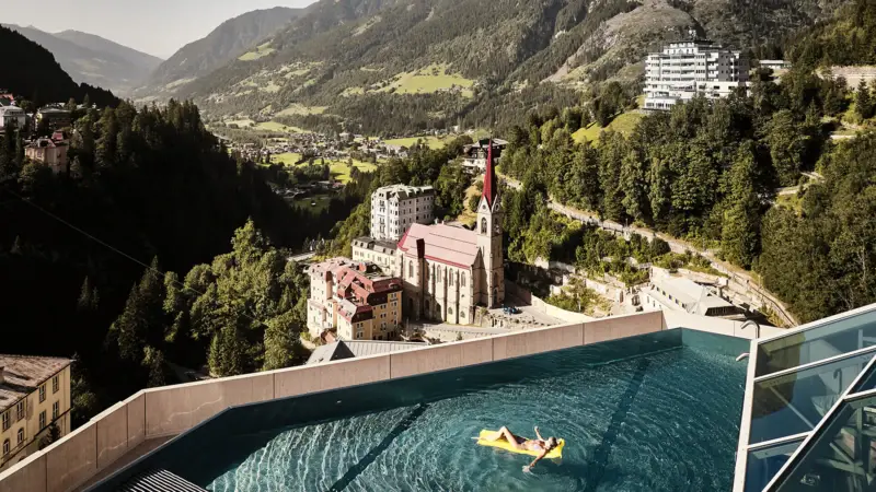 Pool Urban Nature Badeschloss Bad Gastein Eine Frau schwimmt auf einer gelben Luftmatratze auf dem Rooftop-Pool vom Urban Nature Badeschloss Bad gastein mit Blick in das Tal und die Berglandschaft.