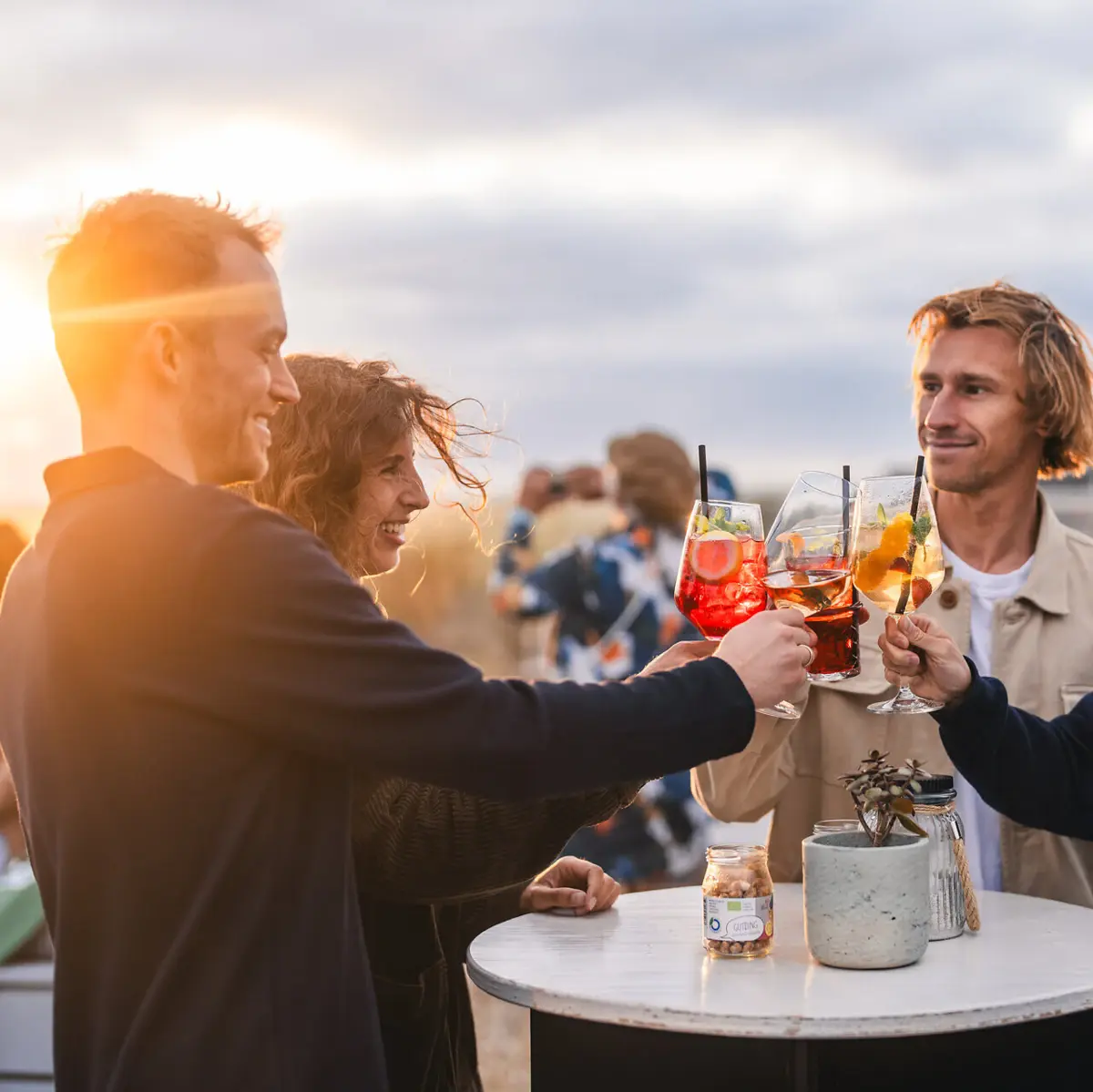 Drinks Scraper’s Club Eine Gruppe von Menschen steht auf der Rooftop Bar Scraper's Club St. Peter-Ording mit Getränken in der Hand und stößt beim Sonnenuntergang an.