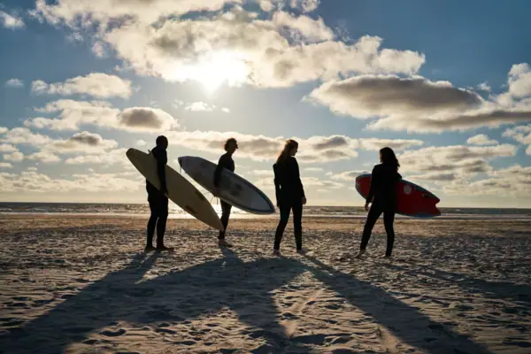 Surfen St. Peter-Ording Vier Menschen stehen an einem Strand und tragen Surfbretter unter dem Arm bei Sonnenschein.