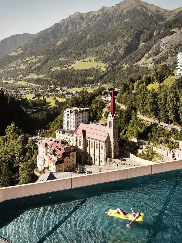 Rooftop Infinity Pool im Badeschloss Bad Gastein Infinity Pool mit Ausblick auf Bad Gastein und die Alpenlandschaft – Frau entspannt auf gelber Luftmatratze mit Blick auf Kirche und historische Gebäude