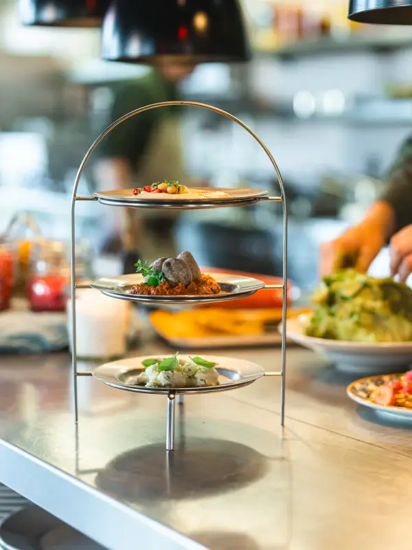 Dips Auntie Clara Eine Etagere mit drei Dips steht auf einer Küchen-Arbeitsplatte und im Hintergrund arbeitet ein Koch.