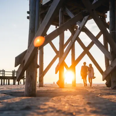 St. Peter-Ording Strandspaziergang Zwei Menschen laufen Arm in Arm beim Sonnenuntergang am Strand von St. Peter Ording zwischen den Pfahlbauten entlang.