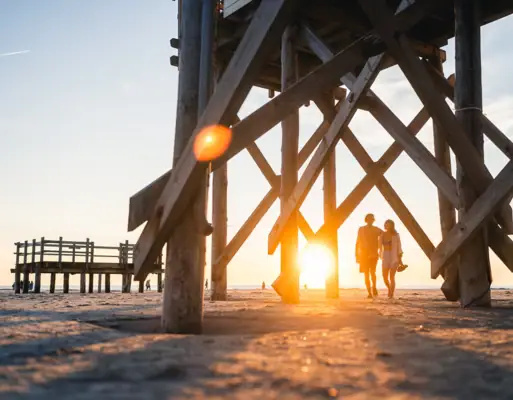 Strand St. Peter-Ording Zwei Menschen laufen Arm in Arm beim Sonnenuntergang am Strand von St. Peter Ording zwischen den Pfahlbauten entlang.