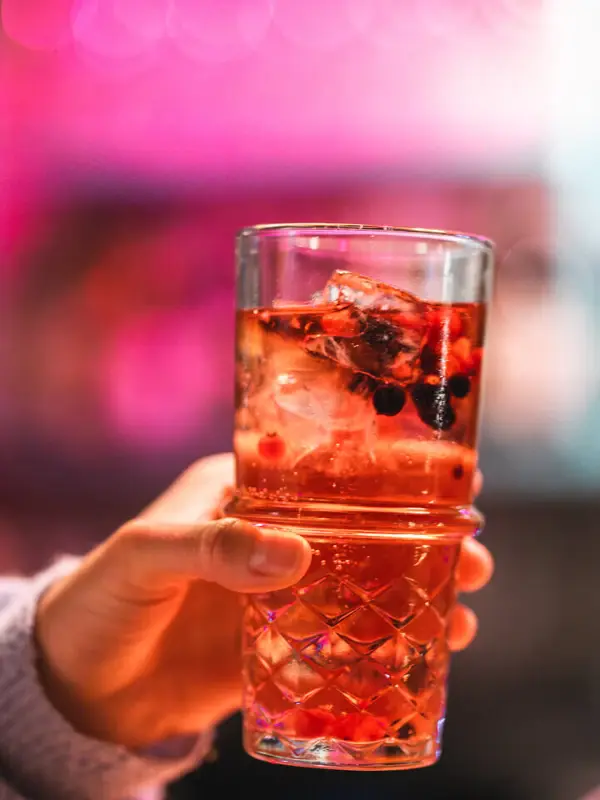 Dink Drifters Hang Out Eine Hand hält einen roten Drink in einem Glas mit Eiswürfeln und Beeren.