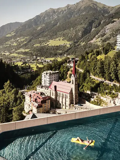 Schwimmen with a view Infinity Pool mit Ausblick auf Bad Gastein und die Alpenlandschaft – Frau entspannt auf gelber Luftmatratze mit Blick auf Kirche und historische Gebäude