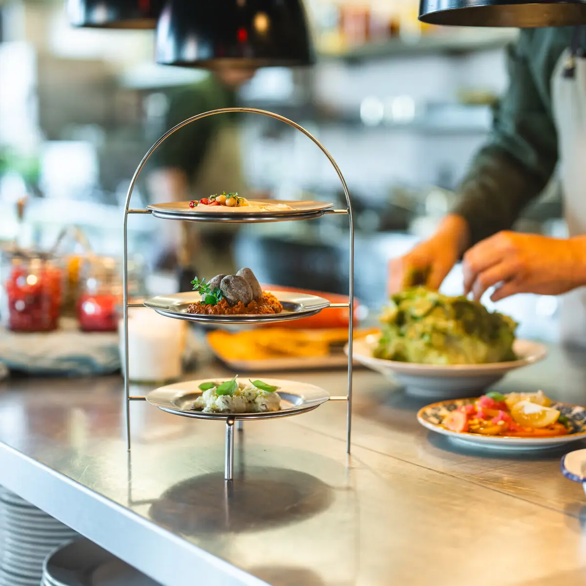 Dips Auntie Clara Eine Etagere mit drei Dips steht auf einer Küchen-Arbeitsplatte und im Hintergrund arbeitet ein Koch.