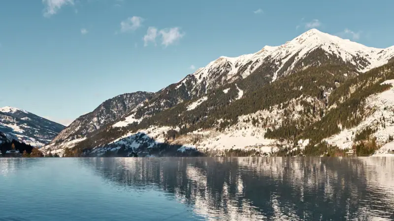 Badeschloss Pool Winter Ein Gewässer mit schneebedeckten Bergen und blauem Himmel.