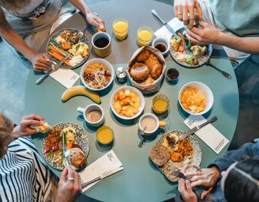 Frühstück Urban Nature St. Peter-Ording Ein runder Frühstückstisch von oben fotografiert, an dem vier Menschen sitzen und Brötchen, Obst und weitere Speisen essen.