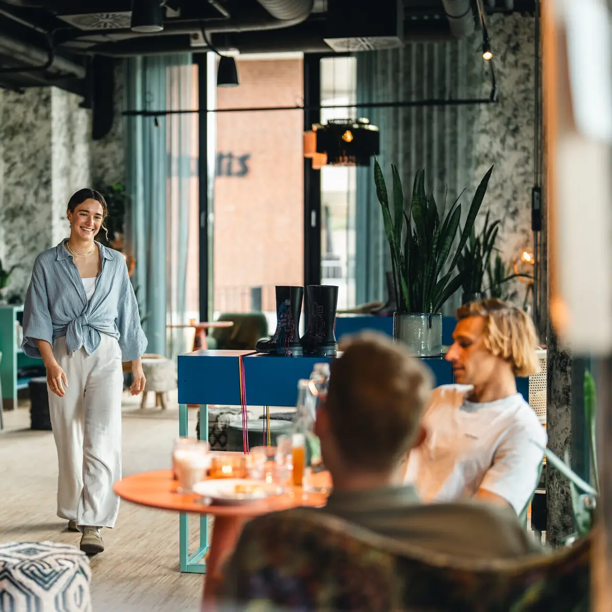 Bar Drifters Hang Out Eine Frau läuft in einer Bar auf einen Tisch zu, an dem schon zwei Männer sitzen und lächelt.