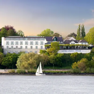 Hotel Louis C. Jacob Hamburg Hotel Louis C. Jacob mit Blick auf die Elbe, Segelboot im Vordergrund, umgeben von grüner Landschaft und Bäumen bei Sonnenuntergang