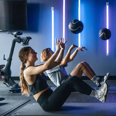 Fitness Urban Nature St. Peter-Ording Zwei Personen sitzen auf einer Matte im Fitnessstudio, halten die Heine und den Oberkörper in der Luft und werfen dabei einen Ball.