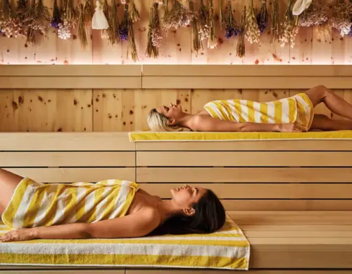 Sauna im Alpine Swim Club Zwei Frauen mit gelb-weiß gestreiften Handtüchern liegen entspannt in einer modernen Sauna mit Holzwänden und getrockneten Blumen an der Decke.