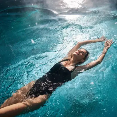 Alpine Swim Club Frau in schwarzem Badeanzug treibt entspannt auf dem Rücken in einem sonnendurchfluteten Pool.