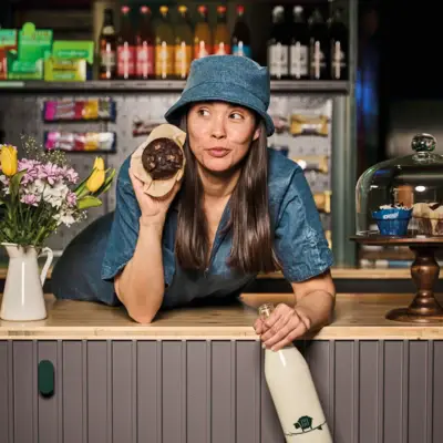 Store & Stuff Eine Frau mit Hut lehnt auf einer Theke und hält einen Muffin in der Hand.