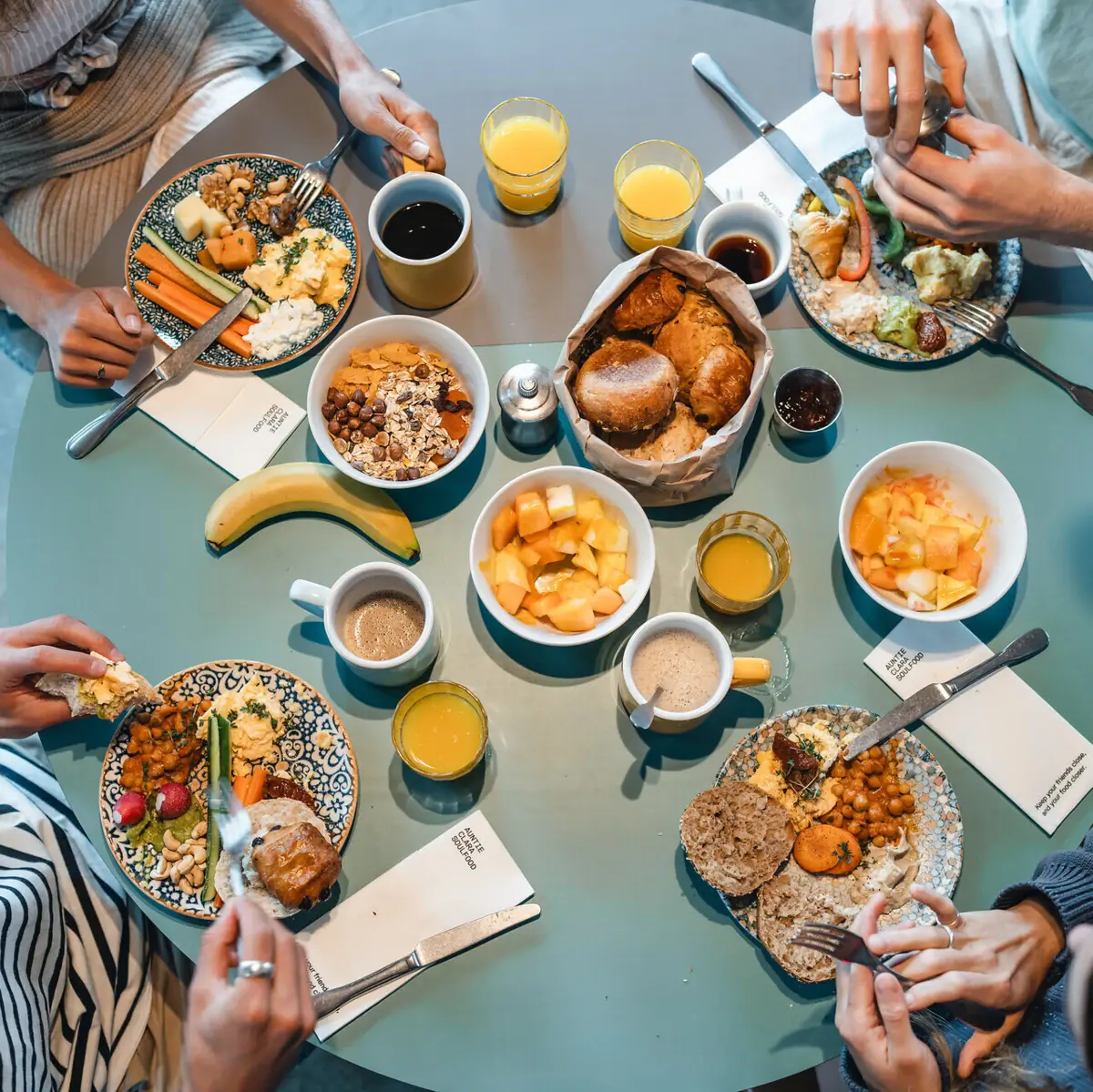 Frühstück Urban Nature St. Peter-Ording Ein runder Frühstückstisch von oben fotografiert, an dem vier Menschen sitzen und Brötchen, Obst und weitere Speisen essen.