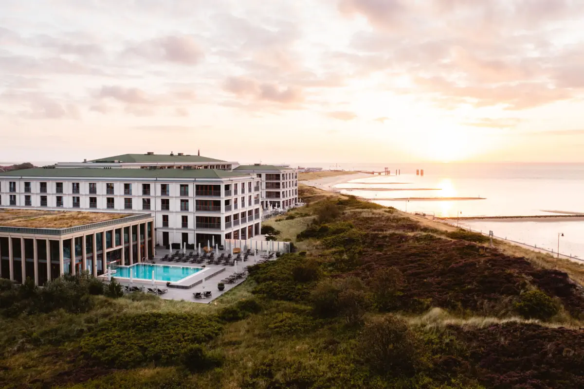 A-ROSA Hotel auf Sylt Modernes Hotel mit Außenpool direkt an der Küste, umgeben von Dünenlandschaft, Sonnenaufgang über der Nordsee im Hintergrund