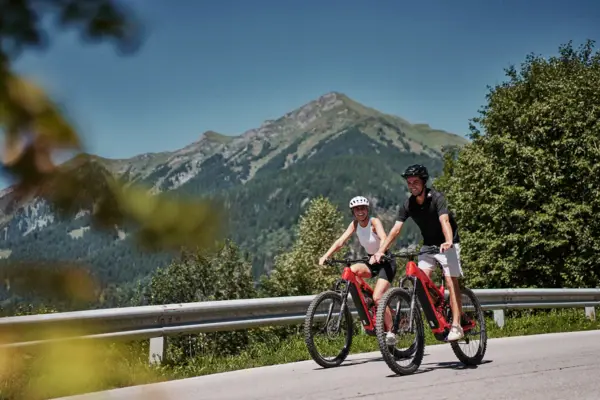 Radtour Bad Gastein Zwei Menschen fahren mit Fahrrädern auf einer Straße entlang und Berge sind im Hintergrund.