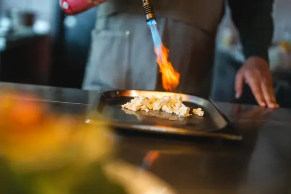 Flambieren Auntie Clara Ein Koch flambiert Blumenkohl mit einem kleinen Flammenwerfer in einer Küche.