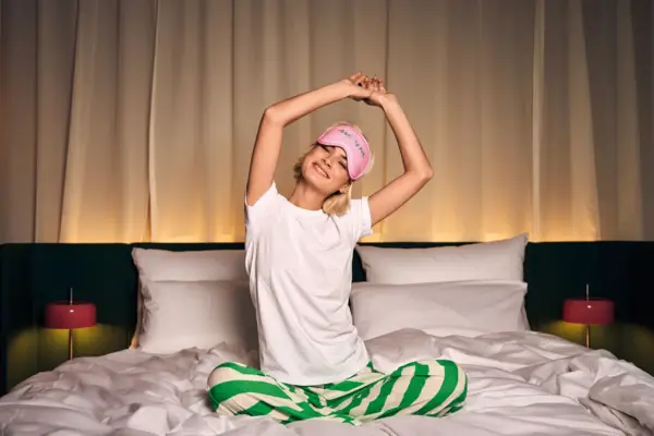 Morgenstimmung im Hotelzimmer im Urban Nature Bad Gastein Junge Frau in grün-weißem Schlafanzug und Schlafmaske streckt sich entspannt auf einem gemütlichen Hotelbett