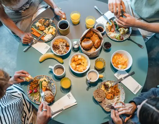 Frühstück Urban Nature St. Peter-Ording Ein runder Frühstückstisch von oben fotografiert, an dem vier Menschen sitzen und Brötchen, Obst und weitere Speisen essen.