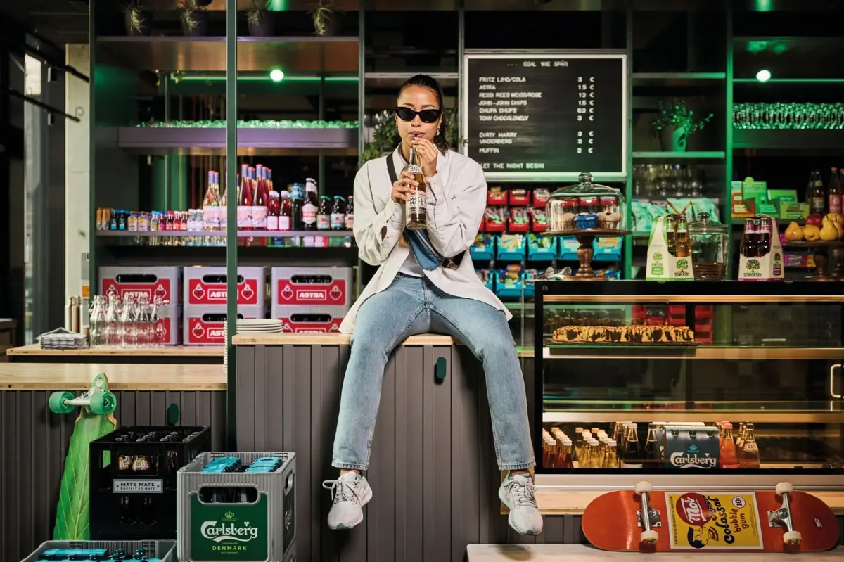 Store & Stiff Kiosk Drink Eine Frau mit Sonnenbrille sitzt auf einer Theke und trinkt ein Getränk aus einer Flasche mit einem Strohhalm.