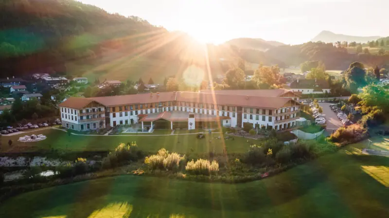 aja Hotel in Ruhpolding Luftaufnahme eines großen Hotels mit weitläufigem Garten in der Alpenlandschaft, Sonnenstrahlen strahlen über die Hügel und den Morgenhimmel