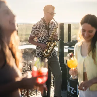 Rooftop Bar St. Peter-Ording Die Rooftopbar Scraper's Club im Urban Nature Hotel St. Peter Ording mit einem Saxophon-Spieler und Menschen, die mit Getränken in der Hand tanzen.