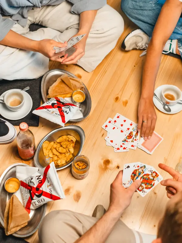 Snacks Drifters Hang Out Mehrere Menschen sitzen im Schneidersitz auf einem Holzuntergrund und spielen zwischen Snacks ein Kartenspiel.