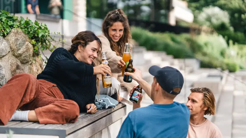 Urban Nature Plaza Eine Gruppe von Menschen sitzt auf einer Kante und hält Getränkeflaschen.